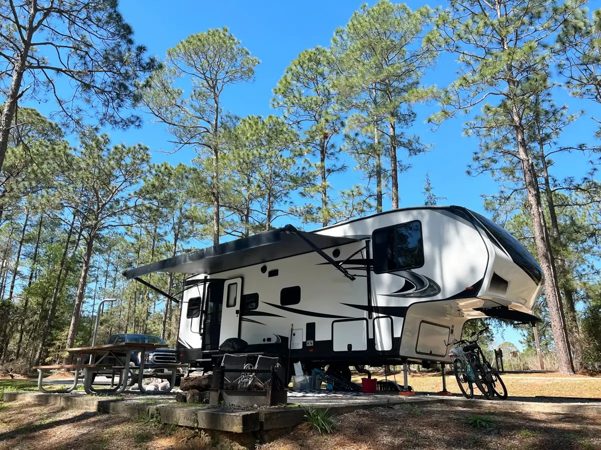 White and black fifth-wheel travel trailer parked at a wooded campsite with an extended awning